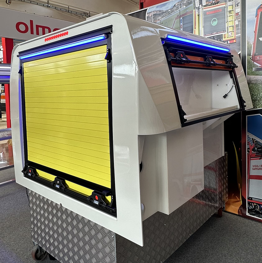 Module d’intervention blanc avec rideau métallique jaune fermé à l’arrière et compartiment latéral ouvert, éclairé par des bandes LED bleues.<br />
